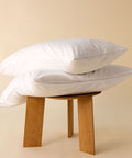 Zwei weiße Kissenschonbezüge dekorativ auf einem Holzhocker präsentiert, mit pflegeleichtem Stoff und schlichtem Design