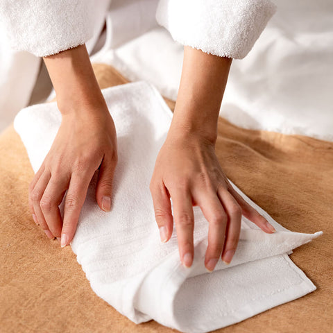 Hände falten ein weißes Handtuch auf einer Holzoberfläche, Symbol für hochwertige Luxushotel-Handtücher und sorgfältige Verarbeitung.