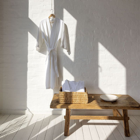 Ein heller, lichterfüllter Raum mit weißer Backsteinwand und Holzdielen, in dem ein weißer Premium Bademantel an einem Kleiderbügel hängt, während ein zweiter zusammengelegter Bademantel in einem Weidenkorb auf einer rustikalen Holzbank liegt.