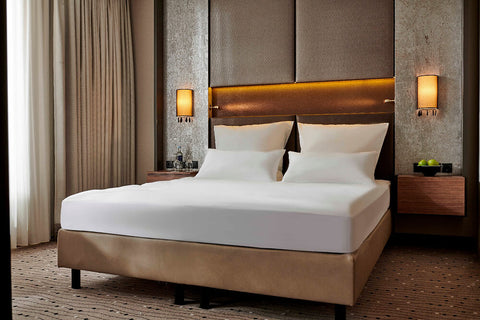 Ein ordentlich gemachtes Doppelbett in einem luxuriösen Hotelzimmer, bezogen mit einem glatten, weißen Premium-Spannbettlaken unter warm beleuchteten Wandlampen und stilvollem Kopfteil.
